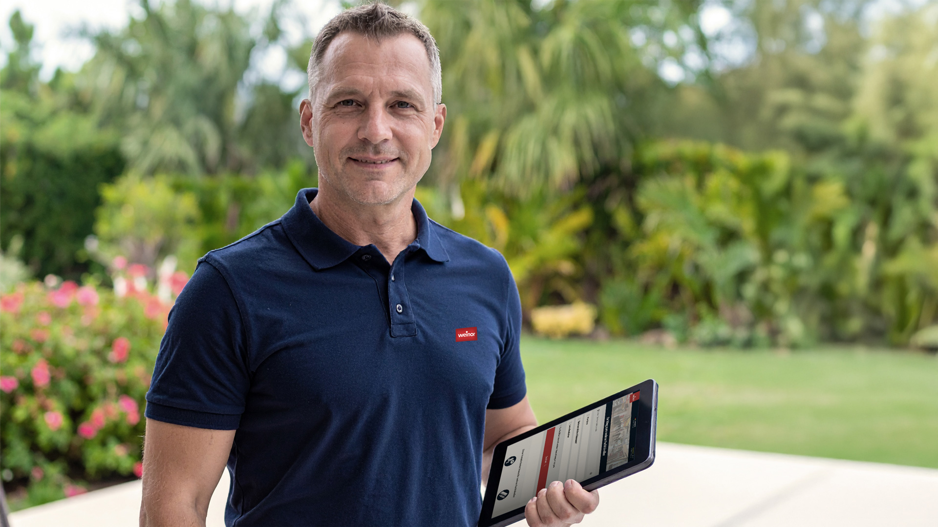 Ein Mann in einem dunkelblauen Shirt hält ein Tablet und steht in einem blühenden Garten.