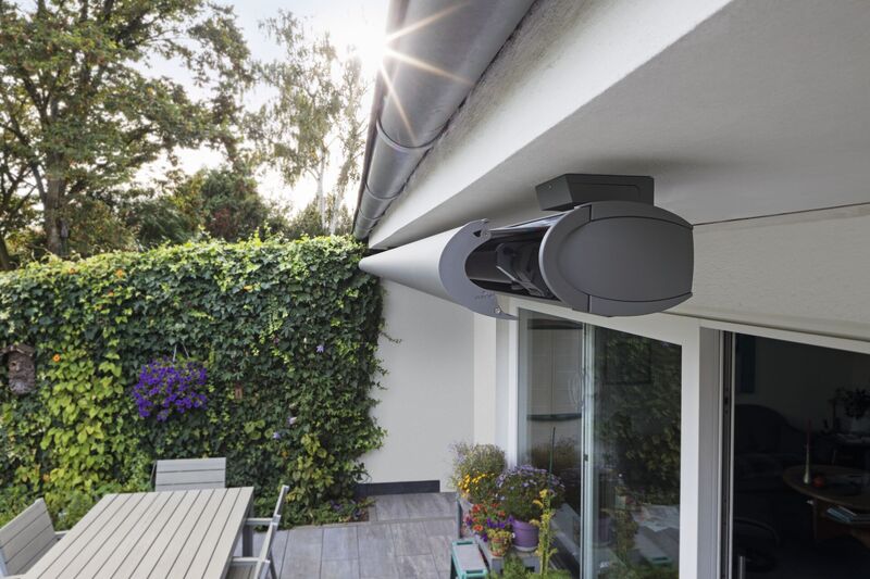 A modern roll-out awning mounted under a roof edge, with greenery in the background and sunlight streaming in.