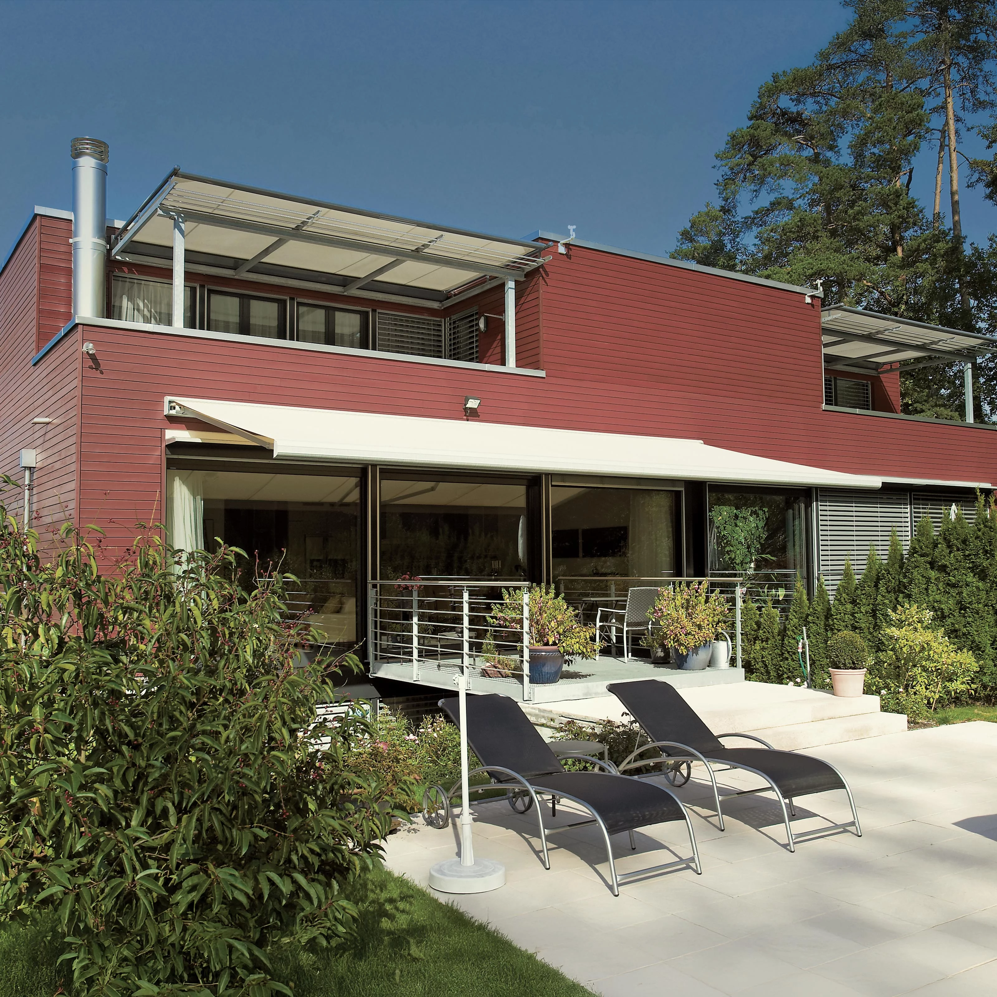 A modern house with a flat roof, large windows, and a terrace featuring seating and green landscaping.