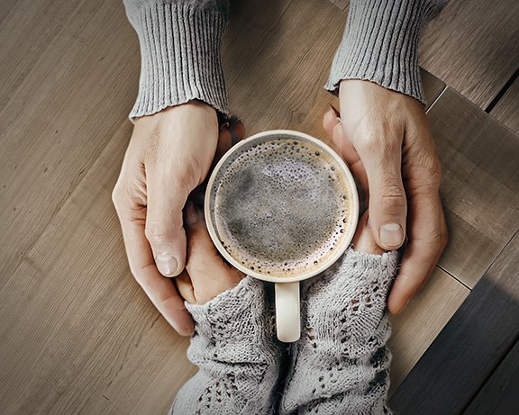 Two hands holding a cup of coffee on a wooden surface, with warm, soft textures present.