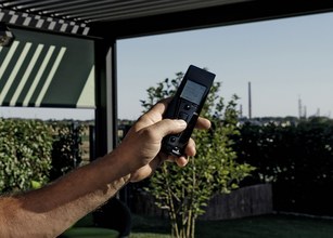 A hand holding a remote control, with greenery and an outdoor setting in the background. 