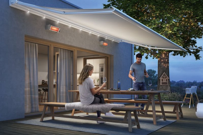 A couple enjoying an evening on a terrace under a retractable awning, with a tree illuminated in the background.