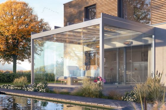 A modern glass structure beside a pond, with greenery and an autumn tree in the background.
