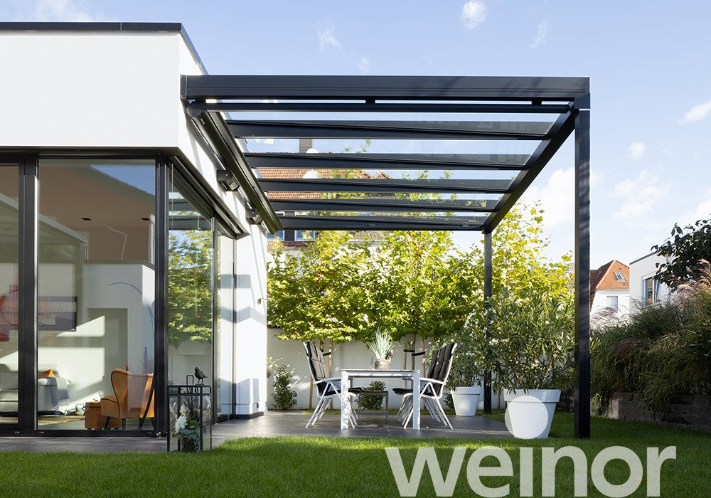 A modern patio with a black pergola, glass roof, white furniture, and plants in a bright setting. 