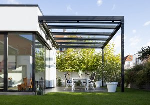 A modern patio with a black pergola, glass roof, white furniture, and plants in a bright setting.