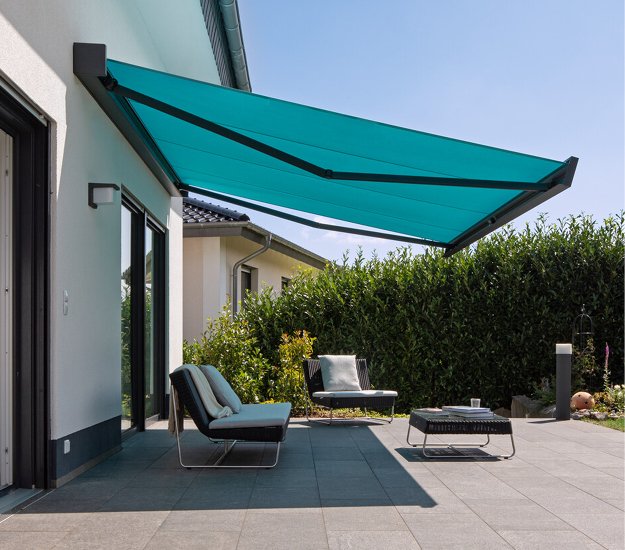 A turquoise awning extends over a patio with two chairs and a table, surrounded by greenery. 