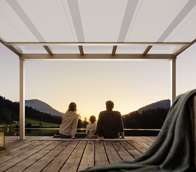 A family sitting on a terrace under a large awning, enjoying a sunset over mountains. 