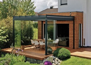A modern terrace with a glass-covered area, wooden elements, and greenery surrounding it. 