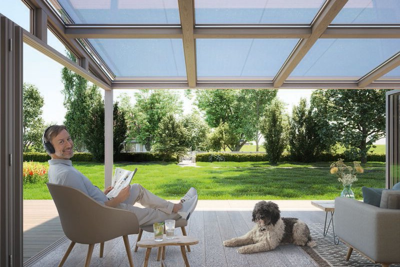 A person sitting in a chair with a dog nearby, inside a modern space with large glass panels and greenery outside. A person sitting in a chair with a dog nearby, inside a modern space with large glass panels and greenery outside.