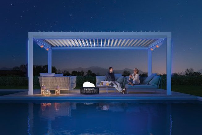 A stylish outdoor seating area under a modern structure, illuminated, beside a calm pool at night.