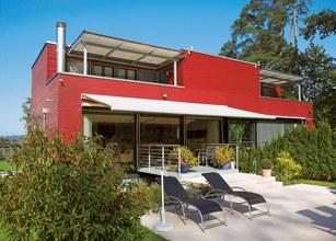 Modern house with a red facade, large glass doors, and a terrace featuring sun loungers and green plants. 