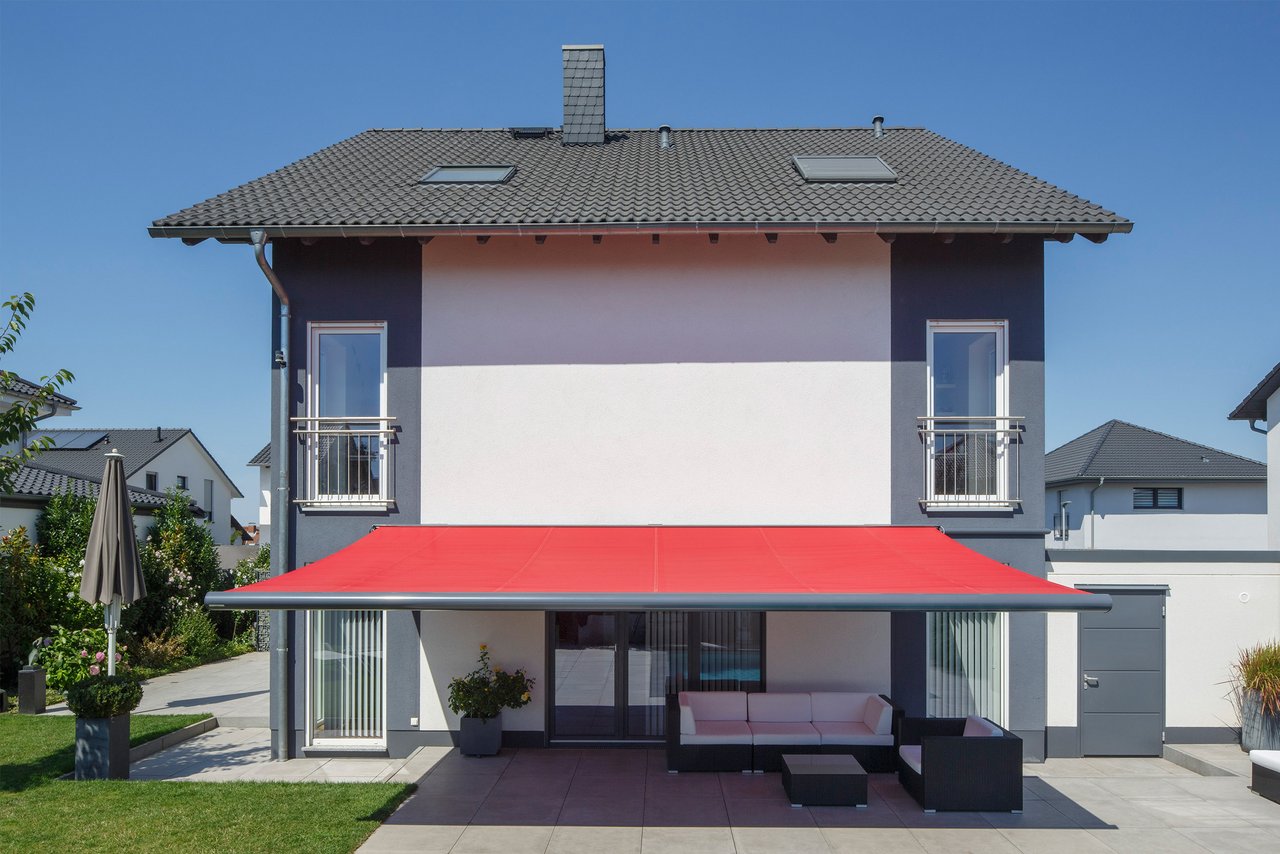 Two-storey house with a pink façade, red awning, and a modern outdoor seating area. Clear blue sky in the background.