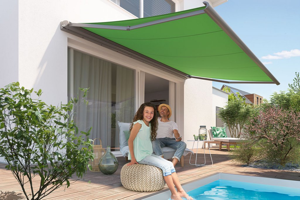 Two people relaxing by a swimming pool under a green awning, with surrounding plants and a wooden deck.