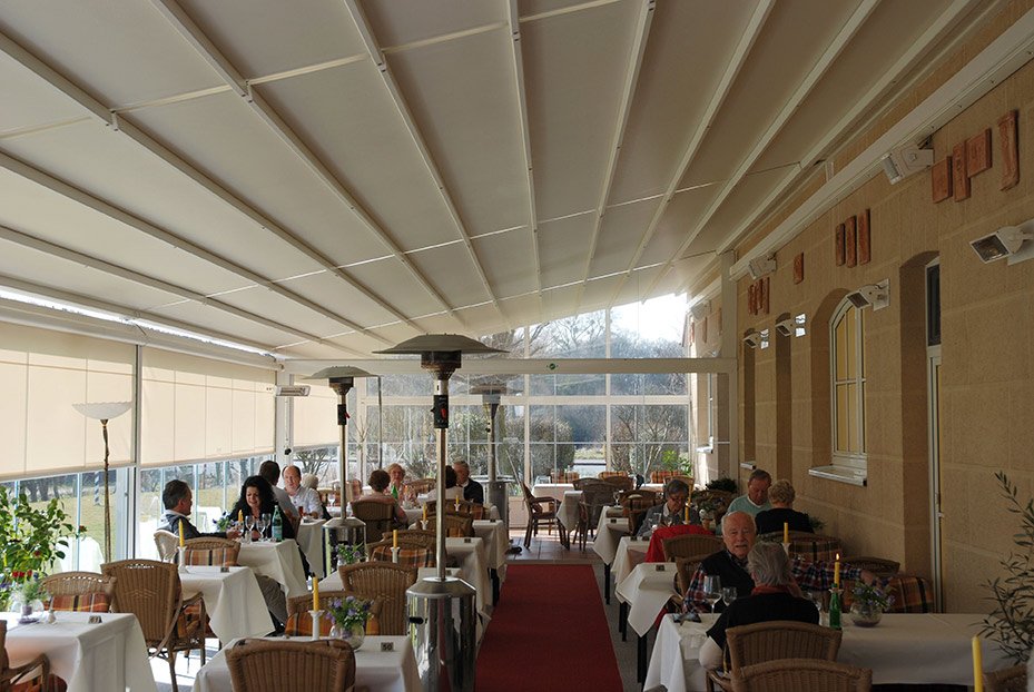 Indoor restaurant with tables, chairs, plants, and a sloped ceiling. Natural light illuminates the space.