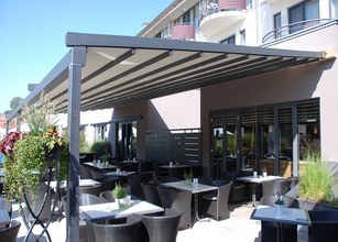 A covered outdoor dining area with dark furniture and greenery, under a sleek grey structure. 