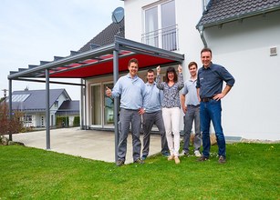 Group of five people posing in front of a terrace with a red awning and a modern house in the background. 