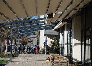 A sunny outdoor space with a covered walkway, children walking, and a decorated tree in the background. 