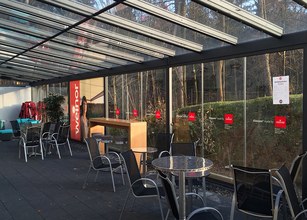 A modern terrace with glass sliding walls, tables, and chairs, surrounded by greenery. 