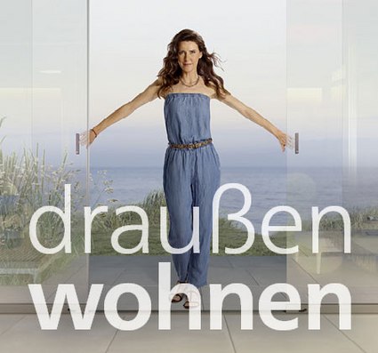 A woman standing with arms outstretched in front of glass doors, with a coastal view in the background.