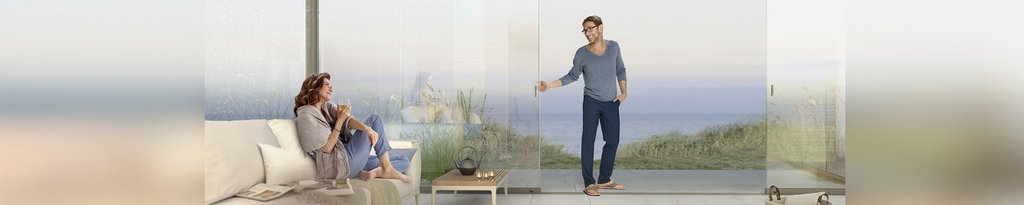 A modern interior with a woman sitting and a man standing by glass sliding panels, overlooking a scenic view.
