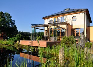 Modern house with a glass terrace overlooking a pond, surrounded by greenery and rocks under a clear blue sky. 