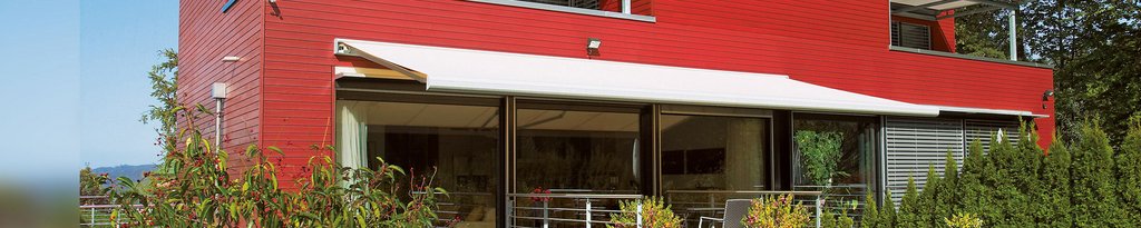 A modern building with a red facade, featuring a white awning and greenery in the foreground.