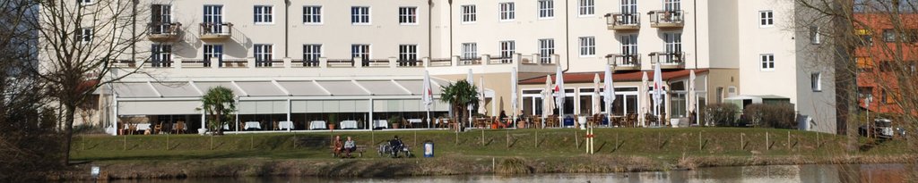 Building facade with large windows, balconies and a terrace near a water body, featuring greenery in the foreground.
