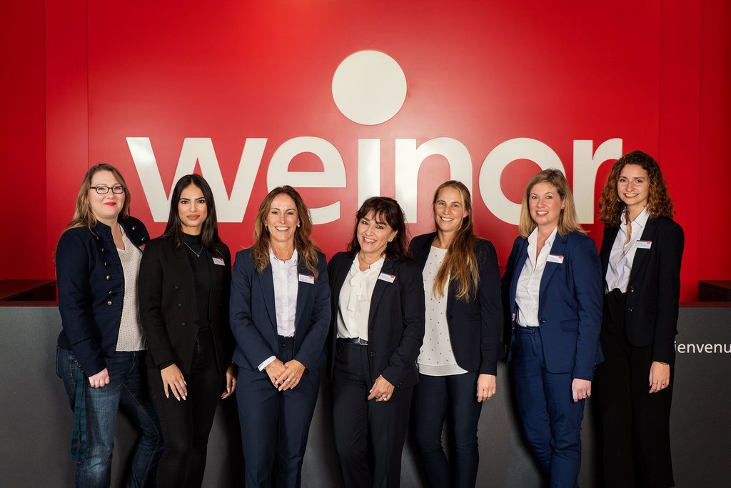 A group of seven women in professional attire stands in front of a red wall with the word "weinor" displayed.