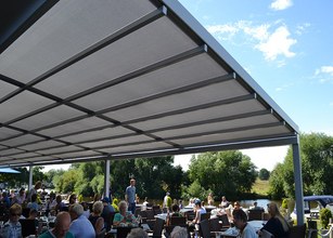 A large overhanging structure provides shade over a busy outdoor terrace by a river. 